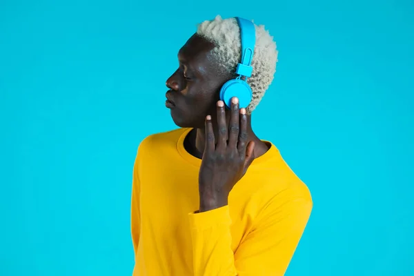 Hombre negro guapo con peinado de moda y auriculares en el estudio sobre fondo azul. Un tipo de traje amarillo. Música, danza, concepto de radio. — Foto de Stock