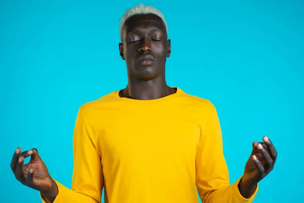 Homem africano calmo em amarelo relaxante, meditando. Ele se acalma, respira profundamente com mudra om no fundo do estúdio azul. Conceito de ioga. — Fotografia de Stock