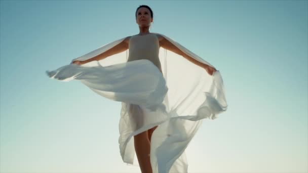 Flexibele vrouwelijke turnster of balletdanser danst met enorme zijden stof fladderend in de wind op de achtergrond van hemel en zon. Concept van tederheid, lichtheid, kunst en talent in de natuur — Stockvideo