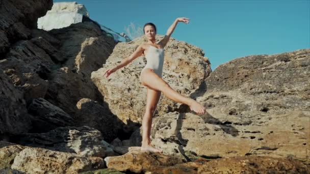 Dansende ballerina tussen rotsen in de buurt van oceaan of zee bij zonsopkomst of zonsondergang. Portret van een flexibele vrouw die in klassieke oefeningen oefent. Concept van tederheid, lichtheid, kunst en talent in de natuur — Stockvideo