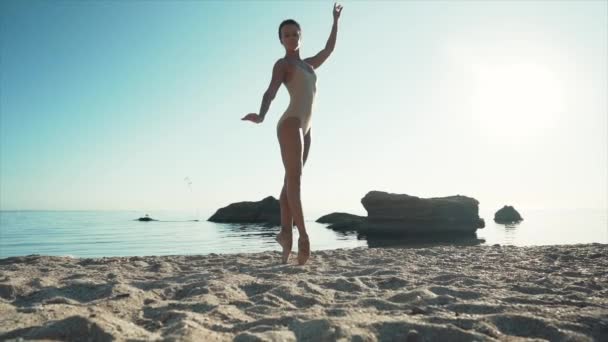 Młoda balerina w beżowym stroju kąpielowym tańcząca balet na morzu lub piaszczystej plaży nad oceanem w porannym świetle. Koncepcja rozciągania, sztuki, piękna natury. Zwolniony ruch. — Wideo stockowe
