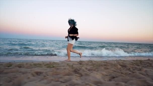 Ładna kobieta z niebieskimi włosami biegnie w pobliżu morza o świcie lub wschodzie słońca, ciesząc się swobodą podczas wakacji letnich, podróży. Piękna szczęśliwa dziewczyna na zewnątrz. Zwolniony ruch. — Wideo stockowe