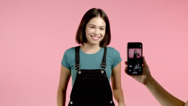 Smilende vlogger kvinne tar opp video av seg selv dansende foran smarttelefon kamera på rosa bakgrunn. Influencer lager rare sosiale medier-klipp – stockvideo