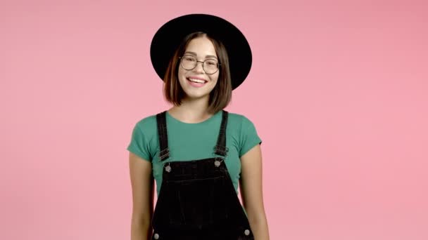Jovem mulher sorridente de chapéu, óculos e olhando em geral para a câmera. Retrato de menina bonita bonito no fundo da parede do estúdio rosa. — Vídeo de Stock