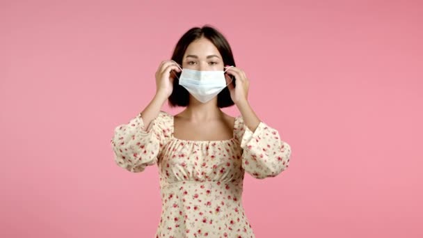 Mujer bonita joven se pone la máscara médica de la cara durante la pandemia de coronavirus. Retrato sobre fondo rosa. Protección con respirador contra brote de COVID-19 — Vídeo de stock