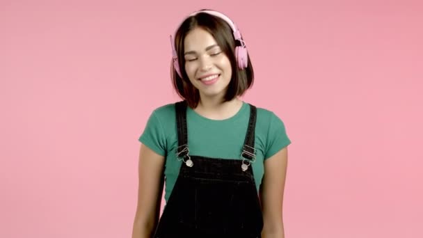ピンクのスタジオの背景にヘッドフォンで音楽を聴きながら踊る魅力的な女性。かわいい女の子の肖像画。ラジオ、幸福、自由、若者の概念. — ストック動画