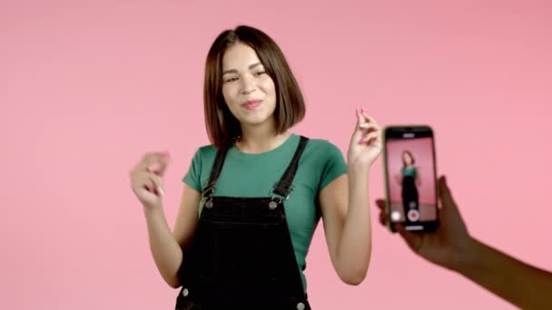 Femme vlogger souriante enregistrant la vidéo d'elle-même dansant devant la caméra du smartphone sur fond rose. Influenceur fait drôle clip de médias sociaux — Video