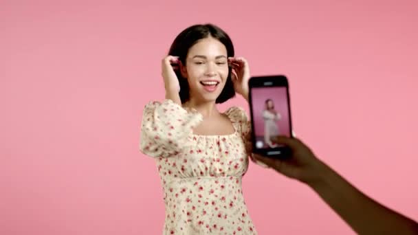 Leende vlogger kvinna inspelning video av sig själv dansa framför smartphone kamera på rosa bakgrund. Influencer gör roliga sociala medier klipp — Stockvideo