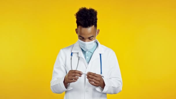 Африканский американский врач в медицинском пальто и маске смотрит на термометр и жестом показывает, что температура не нормальная, пациент болен. Желтый фон студии. — стоковое видео