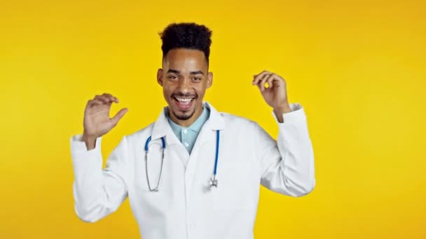 Médecin afro-américain souriant danse drôle, succès et chance au travail. Jeune beau doc homme en manteau médical professionnel blanc est isolé sur fond de studio jaune. — Video