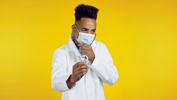 Portrait of serious african doctor in medical coat and mask using stethoscope isolated on yellow studio background. Man points his instrument at camera as if he is listening to ill patient — Stock Video