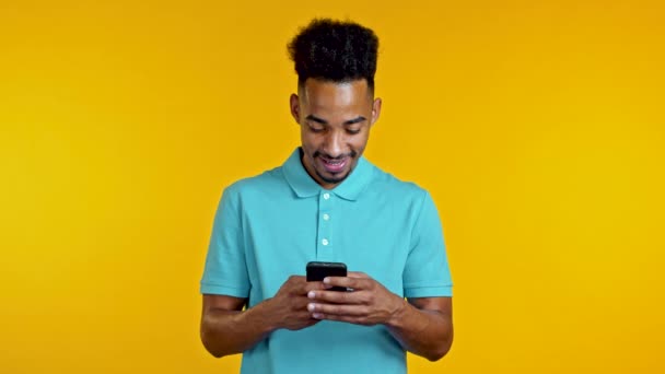 Przystojny Afrykanin używający smartfona na żółtej ścianie studia. Korzystanie z nowoczesnych technologii - aplikacje, portale społecznościowe. — Wideo stockowe