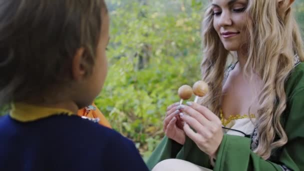 Fee Cosplay junge Frau behandelt kleine Babys Jungen Hobbits mit Bonbons im grünen Wald. Halloween-Konzept, Märchenfiguren, Kinder. — Stockvideo