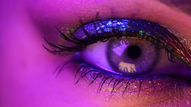 Extreme close-up van het menselijk oog iris onder violet neon licht. Vrouw met mooie make-up, glitter schaduwen. Womens groen oog samentrekken. Schoonheid, cosmetica, nachtclub lifestyle concept. — Stockvideo