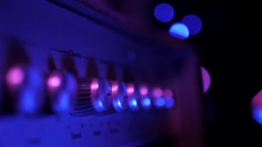 Gitar amplifikatöründeki kontrol panelinin görüntüsü. Taklacılar, düğmeler ve düğmeler. Kadın eli amfinin sesini kısıyor. Mavi ışık, Noel ışığı. Yavaş çekim.