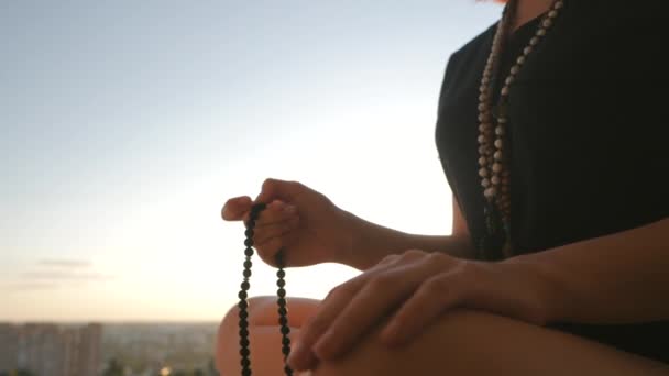 La mano iluminada por las hembras se cierra al atardecer o al amanecer, cuenta las hebras de piedras preciosas de Malas que se usan para mantener la cuenta durante las meditaciones del mantra. La mujer se sienta en la cima de la ciudad. Movimiento lento. — Vídeo de stock