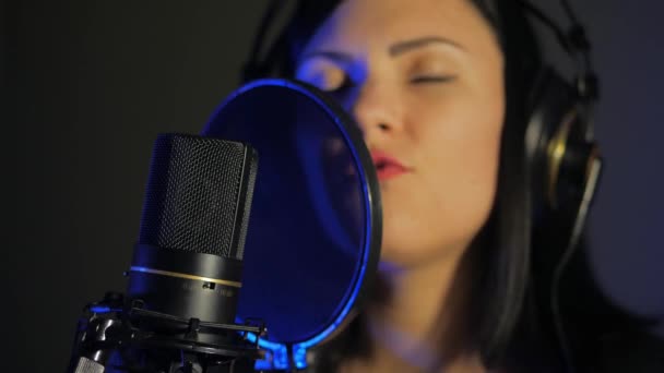 Uma jovem mulher em fones de ouvido canta uma música perto de um microfone em um estúdio de gravação sob luz natural — Vídeo de Stock