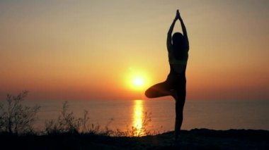 Siluet genç kadın denizin üzerindeki plajda yoga yapıyor. Gün batımında. Fitness, spor, yoga ve sağlıklı yaşam tarzı konsepti. Yavaş çekim.