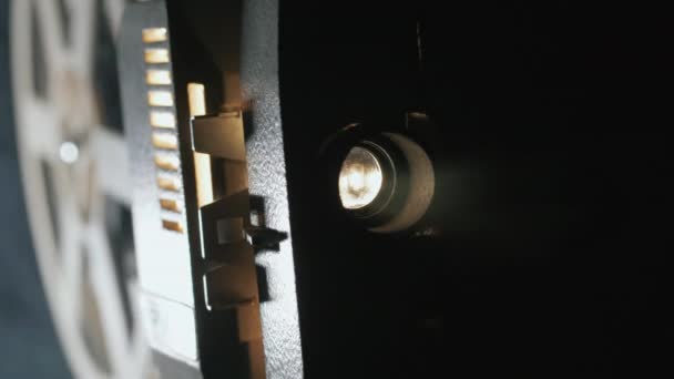 昔ながらのアンティークスーパー8mmフィルムプロジェクターのフロントビュー、暗い部屋に未知のフィルムリールのスタックの横に光のビームを投影. — ストック動画