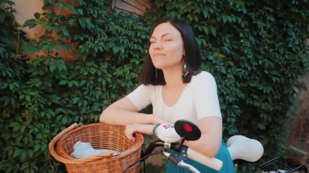 ヴィンテージホワイト自転車で緑豊かな葉の背景に立ってレトロスタイルのスカートを身に着けている短いブルネットの髪を持つ美しい若い女性の肖像。スローモーション. — ストック動画