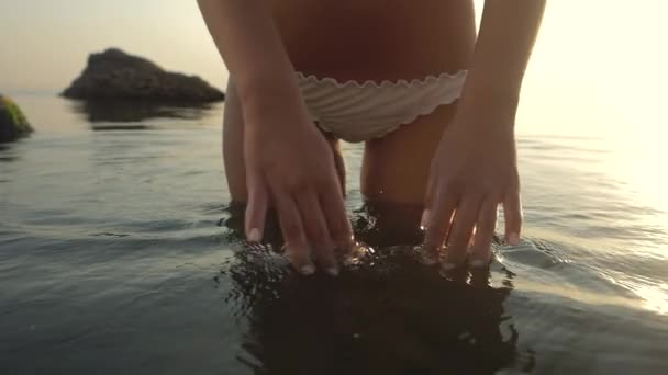 Młoda kobieta w stringach dotykająca wody morskiej w tle wschodu słońca.Powolny ruch.Dziewczyna stojąca na tropikalnej plaży z szczupłym seksownym ciałem w białym bikini cieszącym się letnimi wakacjami w oceanie. — Wideo stockowe