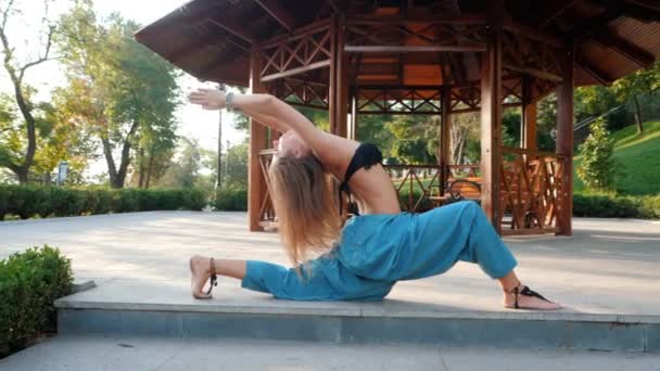 Retrato de yoga matutino de una hermosa joven en pantalones azules y top. Concepto de salud. Chica haciendo asanas en el fondo de la alcoba de madera en el parque en verano. Movimiento lento. — Vídeos de Stock