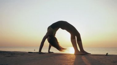 Deniz üzerindeki plajda güneş doğarken vücut kıyafetli genç bir kadın yoga yapıyor. Fitness, spor, yoga ve sağlıklı yaşam tarzı konsepti. Köprü yapan kız, Ağır çekim..