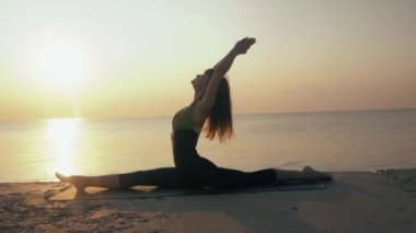 Sabah egzersizi. Tel. Yogacı kadın kalça kaslarını ve kasık bölgesini geriyor. Spor yapan formda sporcu kız gün batımında plajda esneme hareketleri yapıyor. Yavaş çekim.