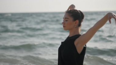 Siyah bluzla dans eden balerin sahnesi ve sabah okyanus ya da deniz kıyısında kot pantolon. Güzel esmer kadın el egzersizi yapıyor. Gimbal atışı. Yavaş çekim.