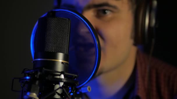 A young man in headphones sings a song near a microphone in a recording studio under natural light — Stock Video