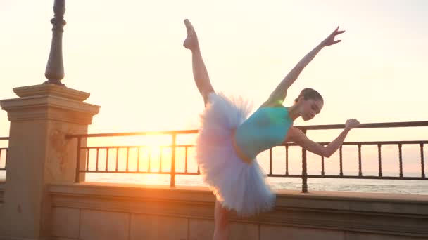 Ballerina in blue ballet tutu and point on embankment above ocean or sea at sunrise. Young beautiful brunette woman practicing stretching and exercises. SLOW MOTION. Close up view, — Stock Video