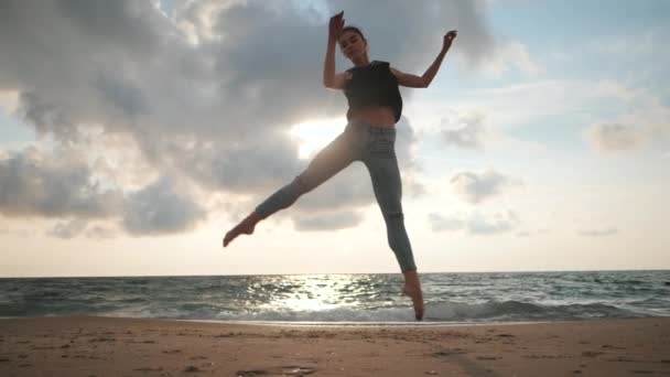 Mujer joven en estilo casual - jeans y top negro haciendo ballet en la playa. Atractivas prácticas de bailarina en saltar sobre la costa arenosa en otoño. Movimiento lento. — Vídeos de Stock