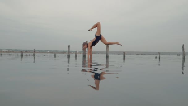 Joven mujer caucásica en traje de baño practicando yoga en agua liman, lago o río. Hermoso reflejo. Asanas complejas, equilibrio. Fitness, deporte, yoga y estilo de vida saludable. Movimiento lento. — Vídeos de Stock