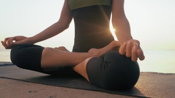 Mujer joven caucásica en traje de cuerpo relajándose practicando yoga en la playa cerca del mar tranquilo, primer plano de las manos, gyan mudra y posición de loto. Fondo del amanecer. Movimiento lento. — Vídeos de Stock