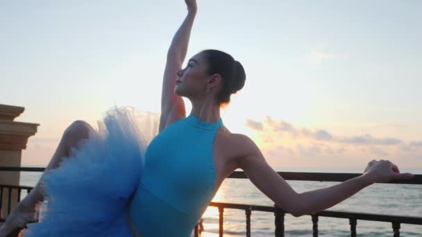 Ballerina di balet biru tutu dan titik pada batu dermaga di atas laut saat matahari terbit. Wanita muda berambut cokelat yang cantik berlatih peregangan dan latihan. SLOW MOTION. Tutup tampilan, — Stok Video