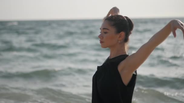 Adegan yang indah menari balerina di atas hitam dan jeans di pantai dekat laut di pagi hari. Wanita berambut cokelat cantik berlatih latihan tangan. Gimbal ditembak. Gerakan lambat. — Stok Video
