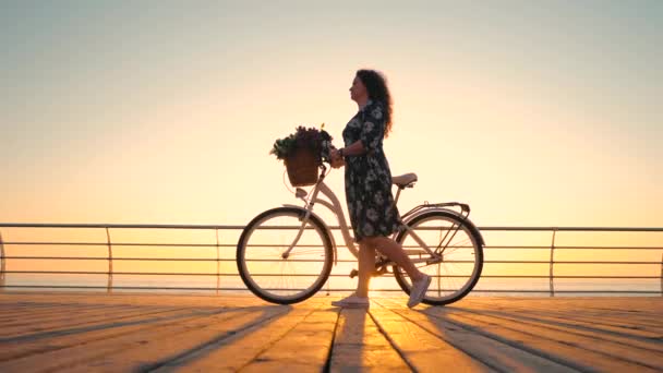 Silhuett av ung attraktiv kvinna i klänning med vintage retro cykel och bukett av blommor promenader på trävall nära havet under soluppgång eller solnedgång. Romantiskt reskoncept. 4k — Stockvideo