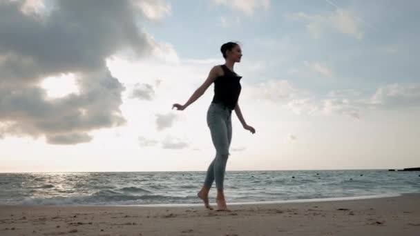 Mulher jovem em estilo casual - jeans e top preto fazendo balé na praia. Bailarina atraente pratica em saltar na costa arenosa no outono. 4k — Vídeo de Stock