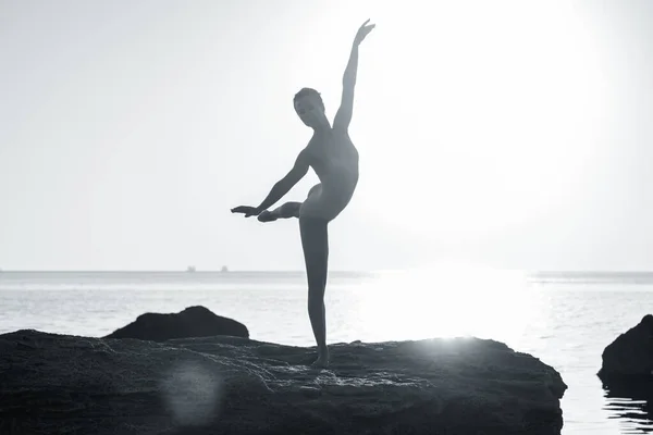 Молодая балерина в бежевом купальнике танцует балет на морском или океанском песчаном пляже при утреннем свете. Концепция растяжения, искусства, красоты природы. — стоковое фото