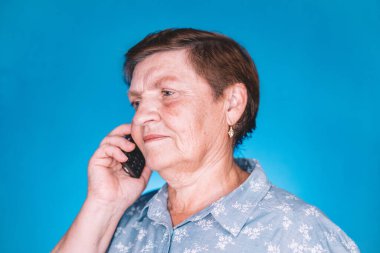 Mavi stüdyo duvarında mobil cihazıyla konuşan yaşlı bir kadın. Büyükannem gülümsüyor. Teknoloji, yaşlılar konsepti.