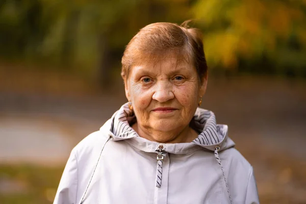 Vackert porträtt av leende äldre kvinna i höstparken. Vit mormor tittar på kameran. — Stockfoto