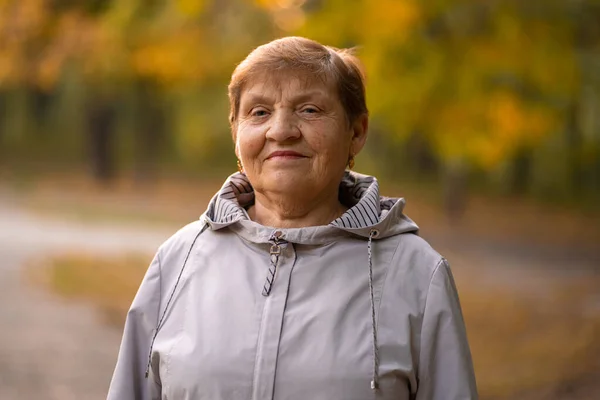 Vackert porträtt av leende äldre kvinna i höstparken. Vit mormor tittar på kameran. — Stockfoto