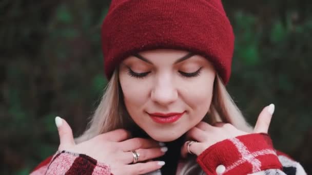 Potret wanita muda cantik di hutan, musim dingin. Gadis yang menarik tersenyum, membetulkan rambut, wanita dengan kemeja trendi dan topi beanie merah. — Stok Video