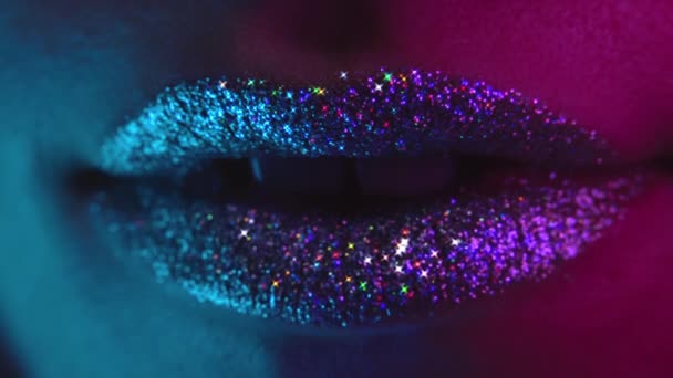 Sexy fashion model with shiny sparkles on lips sending air kiss to camera. Neon ultraviolet colorful light. Macro view of woman with glamorous make-up. Nightlife, night club concept. — Stock Video