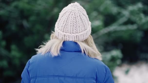 Unrecognizable woman in knitted hat walking in winter forest alone. Young blonde girl in blue puffer jacket. Slow motion. — 图库视频影像