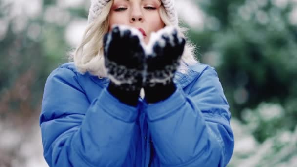 Pretty woman blowing snow from hands with mittens to camera on winter forest background. Portrait of funny girl childishly rejoices first snow. — Stock Video