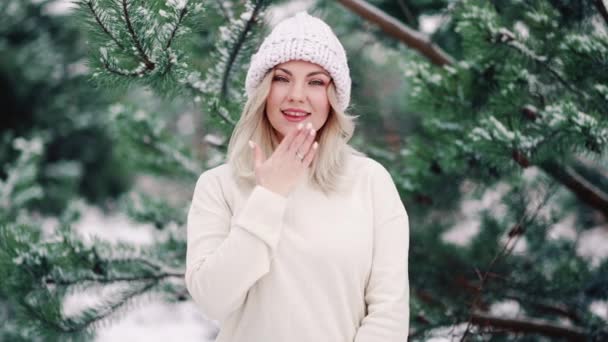 Aantrekkelijke blonde vrouw die een luchtkus stuurt en glimlacht op winterse bosachtergrond. Portret van een jong mooi meisje in een wit pak. Natuur, flirt concept. — Stockvideo