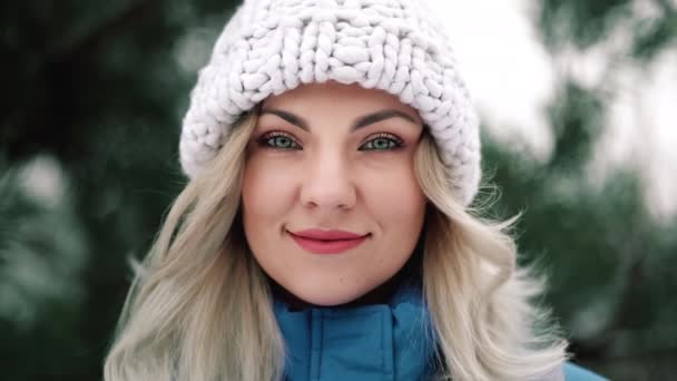 Egy fiatal, csinos nő portréja az erdőben, téli szezonban. Vonzó szőke lány mosolyog, hölgy fehér kötött kalapban. — Stock videók