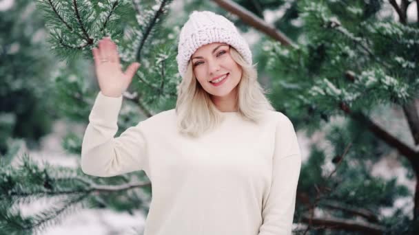 Aantrekkelijke vriendelijke vrouw zwaaiende hand - hallo. Groet, zeg hallo tegen de camera op winterbos achtergrond. Portret van jong mooi meisje in witte outfit. — Stockvideo