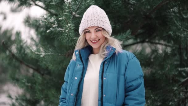 Joven mujer hermosa posando en el bosque durante la temporada de invierno. Atractiva chica rubia tipo sonriente, señora de abrigo azul y sombrero de punto blanco. — Vídeos de Stock